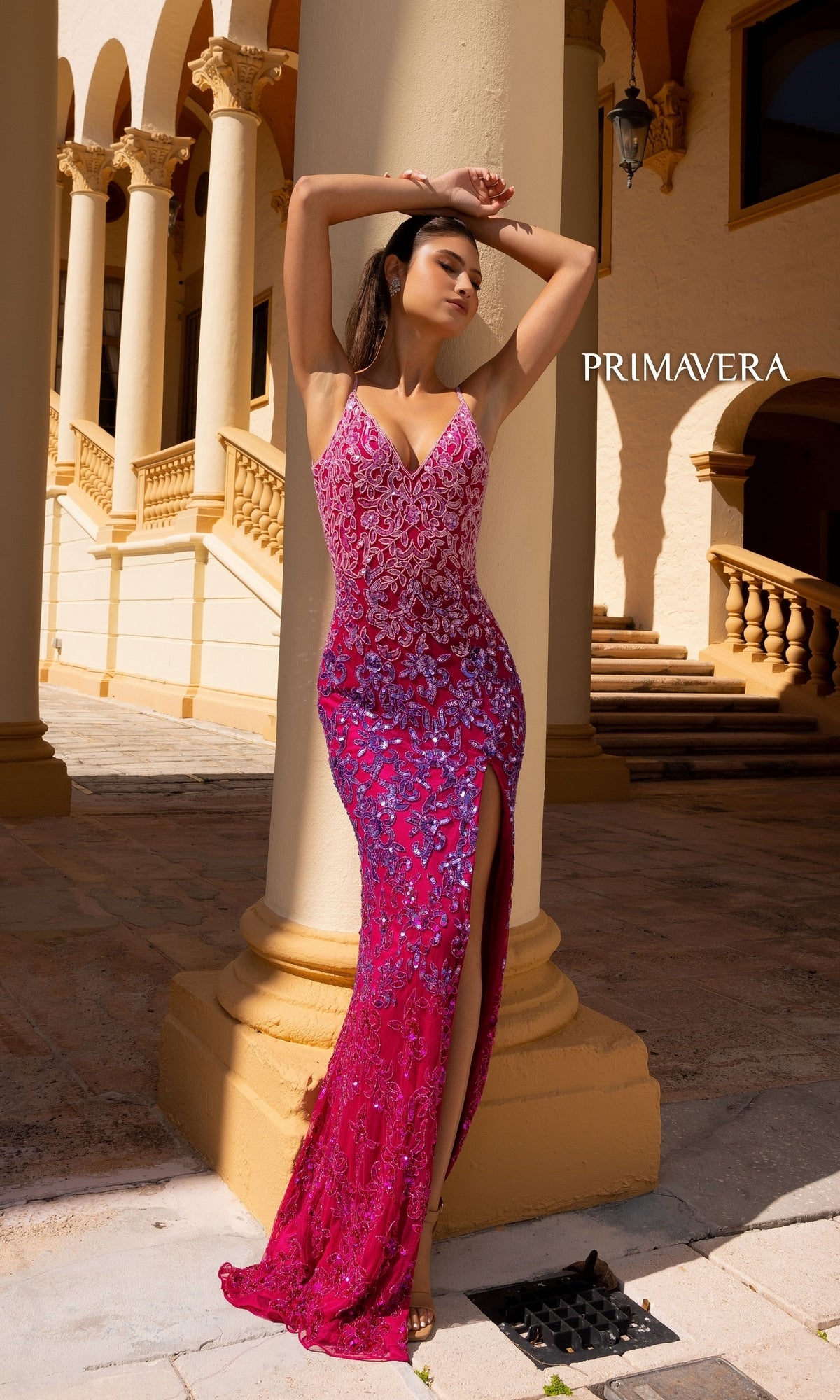A woman poses against a column in front of arched architecture wearing the Primavera Ombre-Beaded Long Formal Gown 4150, a sleeveless pink and purple evening dress with sequins and a thigh-high slit.